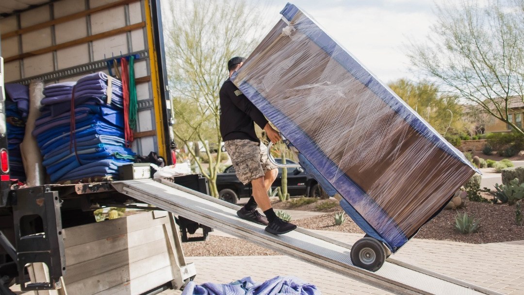 A mover wheeling wrapped furniture down a truck ramp outside a desert landscaped home